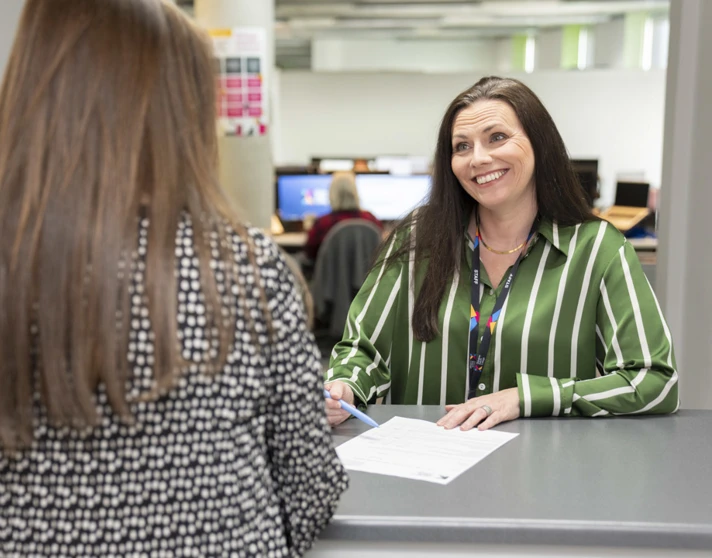 Finance staff member at Glasgow Kelvin College smiling while assisting a student at the service desk, with paperwork on the counter and computers in the background. Finance staff member at Glasgow Kelvin College smiling while assisting a student at the service desk, with paperwork on the counter and computers in the background.